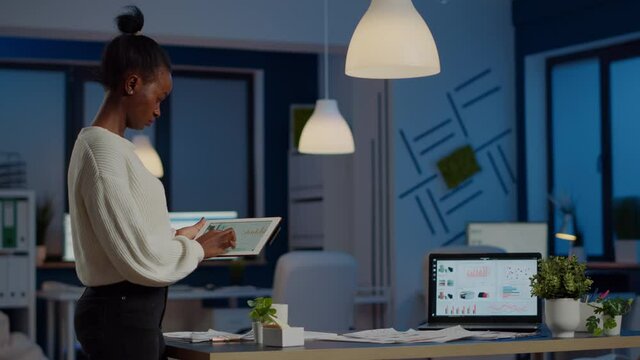 Black woman employee stanging in office and analysing charts during break in business office late at night. Businesswoman using modern technology network wireless overworking in midnight