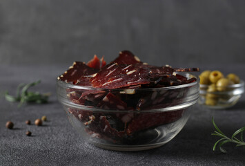 Bowl with jerky, olives, peppers and rosemary on a dark background