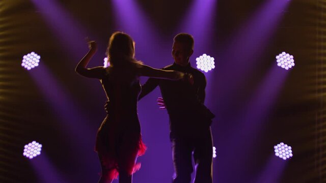 Incendiary rumba performed by ballroom dancing partners. A man in a suit and a woman in a chic short dress dance passionately in a dark studio with bright blue lights. Silhouettes. Close up.