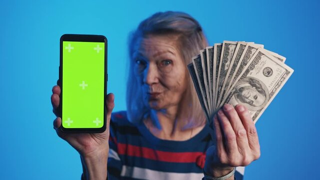 Old woman holding smartphone with green screen and dollar bills. Investment app. High quality 4k footage