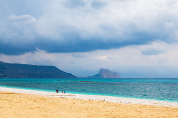 Altea beach, with some people in the distance and with the Ifach rock in the background, on a morning with cloudy skies 