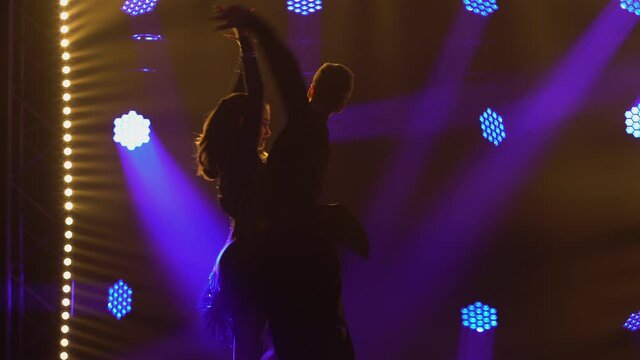 A slender brunette with long hair is dancing the cha-cha-cha with her partner. Silhouette of two people on a dark background with bright spotlights. Close up.