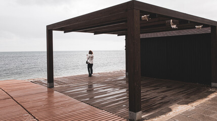 Woman looking to the sea in a rainy day, under a closed beach bar