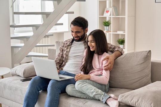 Happy Indian Family Young Father With Teenage Child Daughter Having Fun Using Laptop Computer At Home Watching Tv Movie, Browsing Internet, E Learning, Having Video Call Sitting On Sofa In Living Room