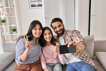 Happy funny indian family with teen child daughter having virtual call meeting on modern cellphone at home. Smiling dad holding cellphone looking at mobile phone taking selfie, recording vlog with kid