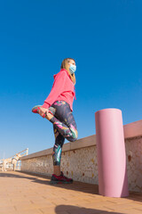 Girl wearing a covid mask and a pink shirt is stretching on the beach while is looking the sea.