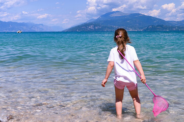 Baby on the beach. A little girl with a net and a bucket plays on the seashore, a large lake. The concept of summer holidays, tourism and travel. Copy space.