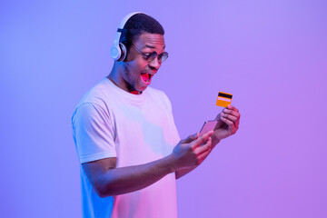 Cashback Concept. Surprised black guy in headphones holding smartphone and credit card