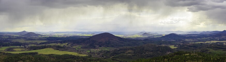 scenic view of rolling hills in the Bohemian Lusatian Mountains with patches of sunlight and storm clouds and rain plumes. Mount Ralsko, Mount Tlustek, Jezevcy vrch and Zeleny vrch can be seen