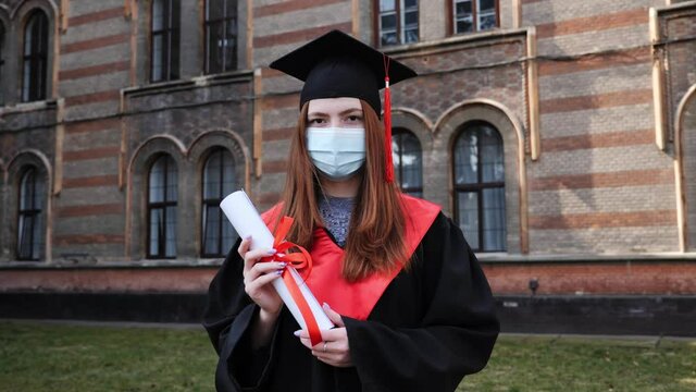 Young graduated girl holding her graduation degree convocation ceremony. Attractive student graduate posing towards the camera in protective mask during coronavirus covid19