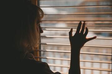 A woman looks out the window through the blinds