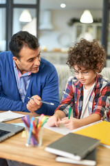 Naklejka premium Best teacher. Loving hispanic father helping his son with homework, explaining task while sitting together at the desk at home