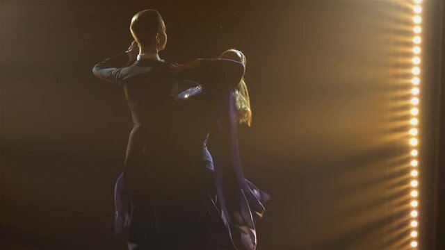 Classical waltz performed by professional ballroom dancers. Silhouettes young man and woman practicing dance steps, performing on floor in dark studio with smoke and spotlight. Close up. Slow motion.