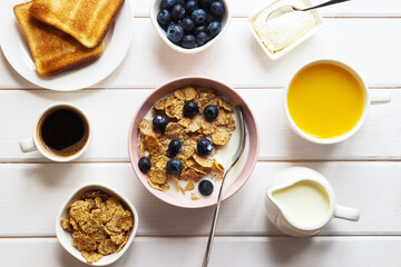 Healthy breakfast of whole wheat flakes with milk, blueberries, cup of coffee, orange juice and toast on white wooden table. Top view, flat lay
