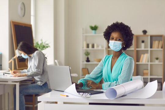 Return to workplace after coronavirus pandemic lockdown. Portrait of black woman in face mask working on laptop in studio office. Architect or designer using computer sitting at desk with paper rolls
