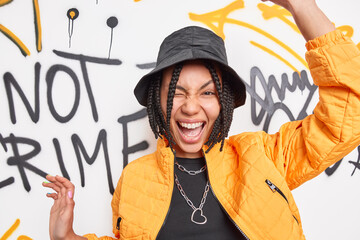 Urban style lifestyle youth and emotions concept. Positive carefree hipster girl has dreadlocks raises arms winks eyes wears black hat and fashionable orange jacket poses against graffiti wall. © WHstudio Leushin N