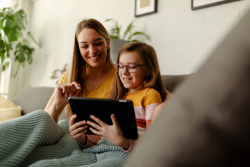 Mom with her daughter is sitting on the sofa and using tablet computer.