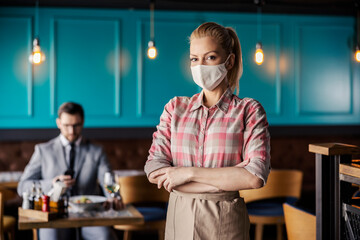 Fototapeta na wymiar Portrait of a female waitress standing in a modern indoor restaurant with her arms crossed. Blonde girl in a plaid shirt and beige apron wears a protective face mask. In the background is a male guest