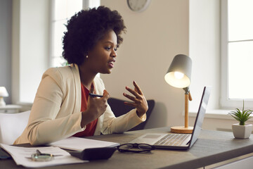 Young african american woman having conference video call using laptop talking to coworker online audience sitting at office desk in evening. Consultation, webinar, tutoring on internet, telecommuting