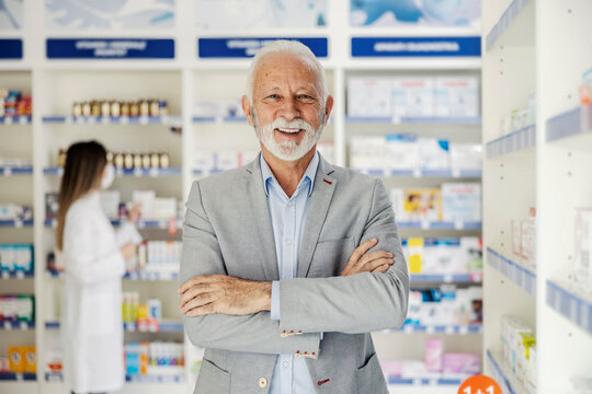 Mature male with arms crossed stands in a pharmacy. Portrait of a man working in a pharmacy in an elegant suit takes a positive attitude towards patients and customers. Woman blurred in the background
