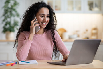 Remote Career. Young Happy Female Freelancer Using Cellphone And Laptop In Kitchen