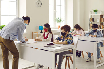 Schools following coronavirus precautions. Children wearing medical face masks in class. Teacher takes, checks and corrects student's test paper while kids are working sitting at desks in classroom