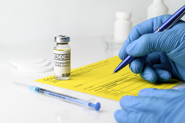 Coronavirus vaccination theme. Doctor's hands in medical gloves filling out the vaccination record card, a vial with covid-19 vaccine and syringe on a white table, close up