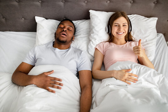 Happy young woman wearing headphones and showing thumb up gesture while her husband snoring on bed, top view