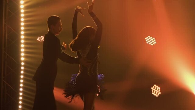 A slender brunette with long hair is dancing the cha-cha-cha with her partner. Silhouette of two people on a dark background with bright colored spotlights. Close up. Slow motion.