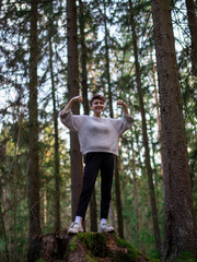 A young beautiful teenage girl smiles and stands in a forest with tall trees on a stump in full growth and shows a pose of strength. The concept of a charge of energy from nature.