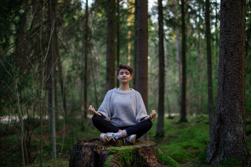 Obraz premium A dark-skinned teenage girl with a brunette with short hair is doing yoga in a green forest on a stump. The lotus position. Casual clothing. The concept of a healthy lifestyle and unity with nature.
