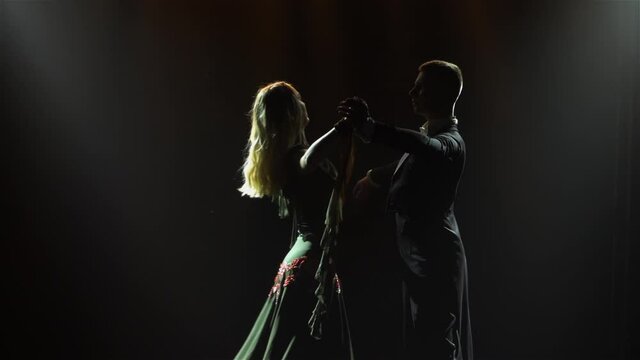 In dark among smoke, silhouette of couple is seen dancing elements of a waltz. Man invites his partner woman to dance. Pair of dancers are illuminated by beams of bright light. Close up. Slow motion.