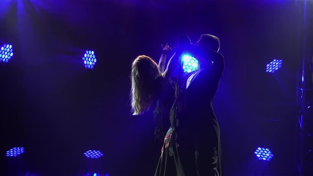 Silhouette of men and women in elegant outfits waltzing in a dark studio. A romantic couple is dancing a classic waltz choreography against the backdrop of blue lights. Close up. Slow motion.