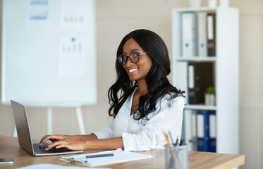 Happy young black businesswoman in glasses sitting at desk with open laptop at company office