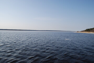 Obraz premium The undulating surface of a wide river under the spring sun. Blue water surface with small waves under a light blue sky. A path from the sun is reflected in the water. In the distance, a dark strip of