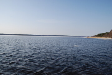 Obraz premium The undulating surface of a wide river under the spring sun. Blue water surface with small waves under a light blue sky. A path from the sun is reflected in the water. In the distance, a dark strip of