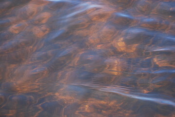 The surface of clear water with small waves. A close-up of a clear river water with a brown bottom from the sand. The reflections of the sun's rays paint the bottom of the river in golden tones.