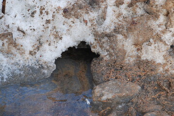 A deep puddle in the snow with melted water. A wide and deep puddle formed in the snow. A puddle flows in a stream into a small hole in the white snow. The snow is white with brown from mud and sand.