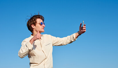 A Spanish white man with long hair, purple glasses and a beige shirt taking a selfie while making a thumbs up sign on clear blue sky background