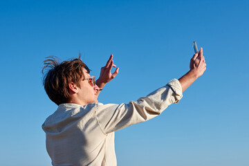 Fototapeta na wymiar A Spanish white man with glasses and a beige shirt taking a selfie and making a victory sign on clear blue sky background