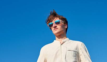 A Spanish white man with long hair, purple glasses and a beige shirt staring into the distance on clear blue sky background