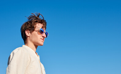 A Spanish white man with long hair, purple glasses and a beige shirt glaring to the side on clear blue sky background