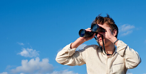 A Spanish white man in a beige shirt looking through binoculars on cloudy sky background