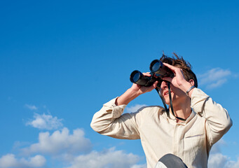 A Spanish white man in a beige shirt looking through binoculars on cloudy sky background