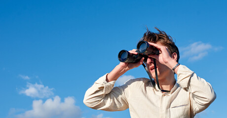 A Spanish white man in a beige shirt looking through binoculars on cloudy sky background