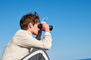 A Spanish white man in a beige shirt looking through binoculars and holding a mask on clear blue sky background