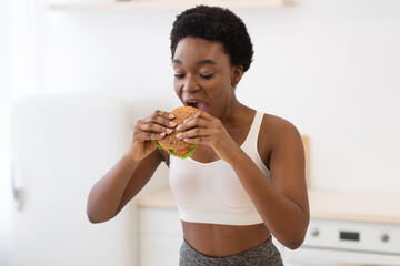Hungry Black Female Eating Big Burger Standing In Modern Kitchen