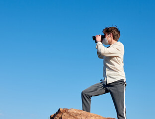 A shallow focus shot of a young man from Spain in a white shirt observing from the cliff with binoculars during the pandemic