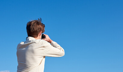 A close up of a young man in a white shirt looking through binoculars on blue space -view from the back