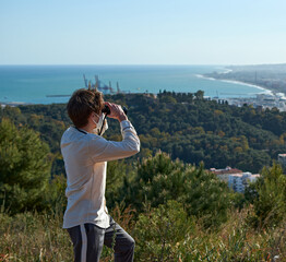 A shallow focus shot of a young man from Spain in a white shirt wearing a mask observing the coastal town through binoculars